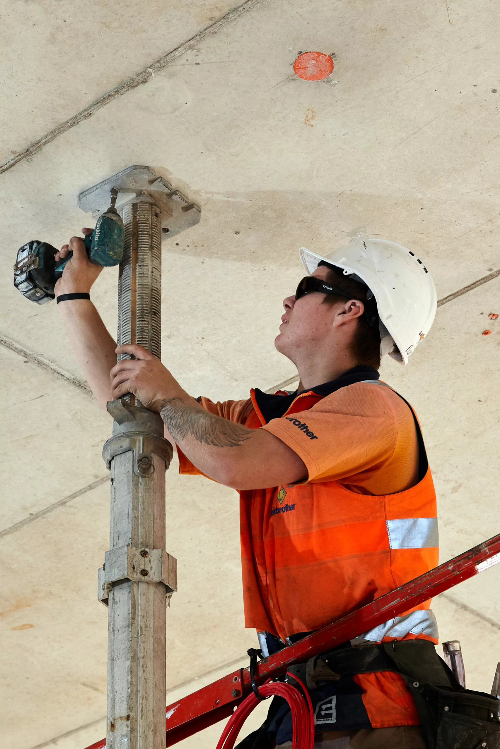 A tradesperson working at height with safety gear drills a screw into a concrete ceiling.