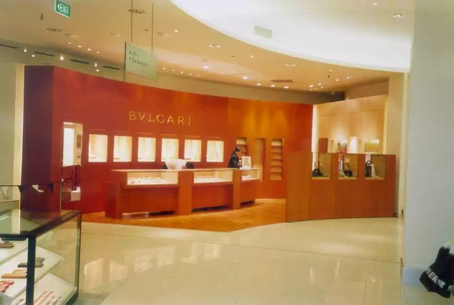 Stylish store attendants work behind glass counters of the Bvlgari high-end fashion and jewelry store. Polished reflective floors are in the foreground.