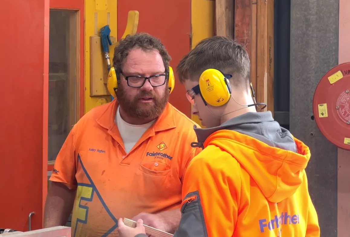 A young Fairbrother apprentice or work experience student wears safety gear and receives instruction from a Joinery master.