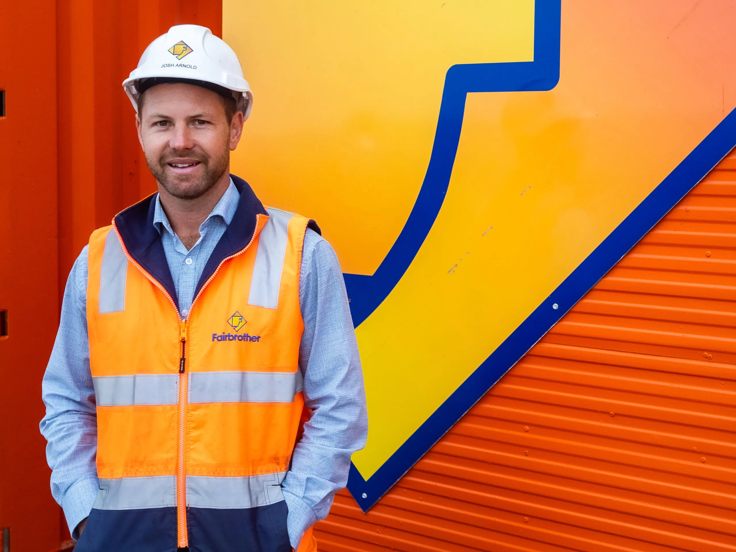 Fairbrother's 2023 Employee of the Year, Josh Arnold dressed in hi vis and a hard hat, stands in front of a Fairbrother site shed with the company logo prominent behind him.